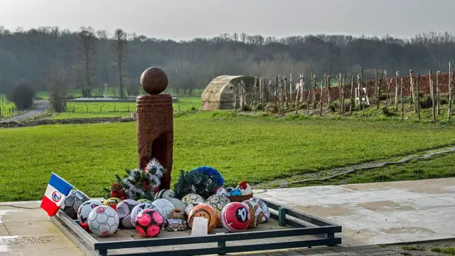 Пам'ятник Різдвяному перемир'ю і футбольному матчу під час нього в Бельгії. Позаду на зеленому полі стоять кілки, які нагадують часи Першої світової війни, а на бетонних плитах стоїть пам'ятник із футбольним м'ячем на верху, на постаменті лежать багато справжніх, кольорових футбольних м'ячв