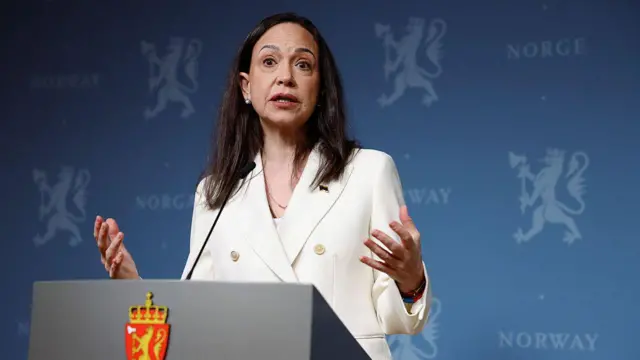 María Corina Machado frente a un podio durante una conferencia de prensa en noruega. Lleva el cabello negro hasta los hombros. Tiene una expresión seria mientras habla. Tiene las manos extendidas hacia los lados. Viste un chaleco blanco. 