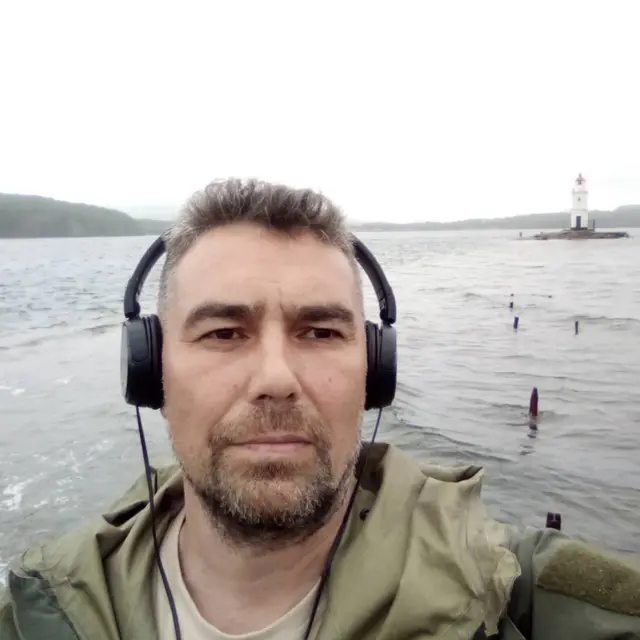 Андрей Акузин в больших черных наушниках фотографирует сам себя на фоне реки и холмов