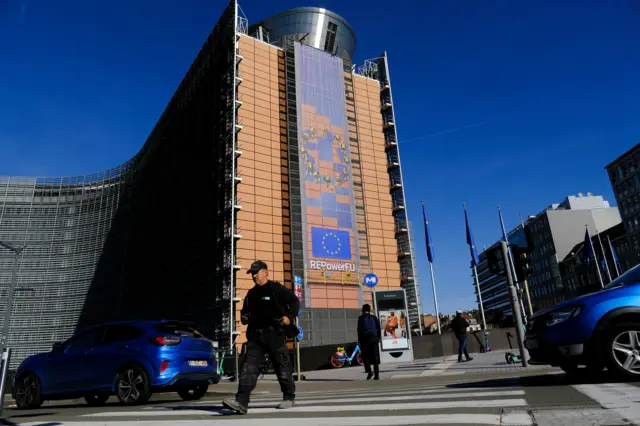 На фоне синего неба стоит многоэтажное здание с флагом Евросоюза и написью REPowerEU. Перед зданием - флаги Евросоюза, идут люди и едут машины.