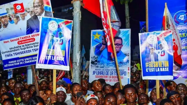 Imbaga y'abantu baje kwakira Tshisekedi i Kisangani, inyuma yabo hari ibyapa biriho ifoto ye biriho n'amagambo yo kumuha ikaze, ku wa gatatu, ku itariki ya 23 Ukwakira (10) mu mwaka wa 2024