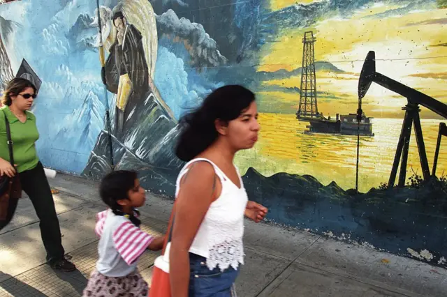 Duas mulheres e uma menina caminham em calçada de Caracas, com um mural pintado no muro ao fundo, retratando uma plataforma de exploração de petróleo, nas cores amarelo e azul