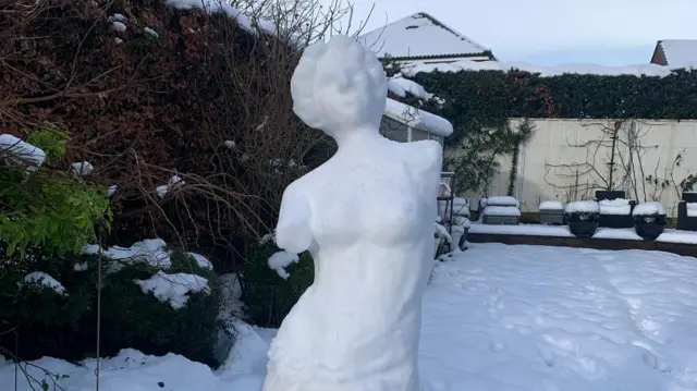 Escultura de estilo griego clásico de una mujer sin brazos, hecha de nieve. Detrás de la escultura se encuentra un jardín suburbano nevado.