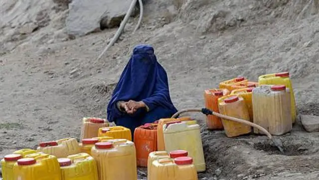 یک زن در حال در کردن بشکه‌های زرد از آب نل. زن در میان بشکه‌ها در حالی نشسته که چادری به سر دارد.