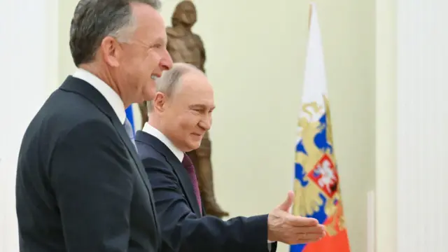 Усміхнені Путін і Віткофф всередині урочистої зали з кремовими стінами. Путін жестом руки показує напрямок. На задньому плані видно російський триколор із гербом, а також бронзову статую.