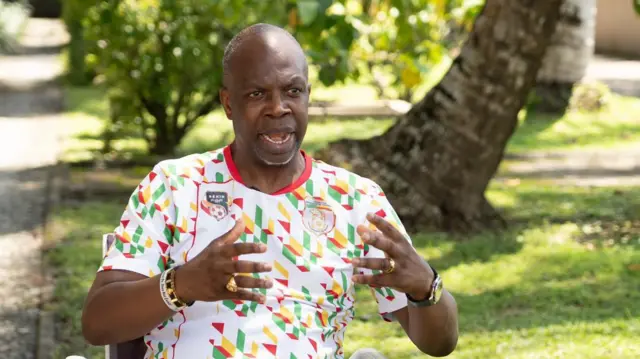 Tunde Adelakun vêtu d'un survêtement du Bénin, assis, la bouche et les mains ouvertes.