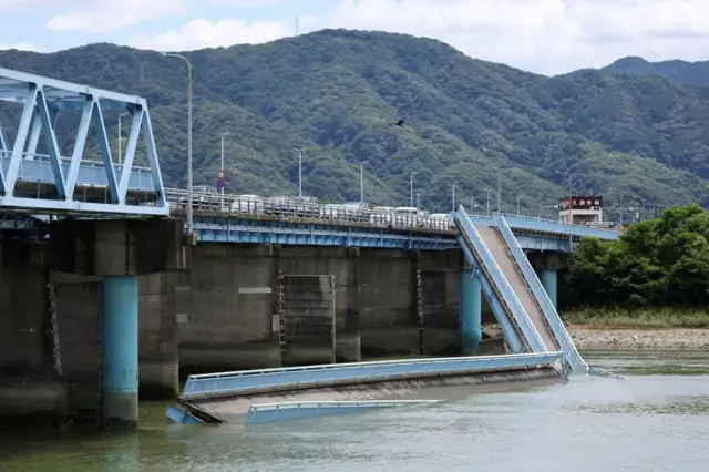 緑に覆われた山を背に、水色の橋が川に架かり、歩道部分の一部が川に落ちている。橋の残る部分には車が連なっている