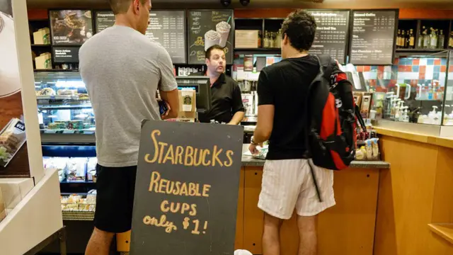 Dois clientes fazendo pedido no caixasoccer bet prouma unidade da Starbucks, onde se vê uma placa oferecendo a vendasoccer bet procopos reutilizáveis por 1 dólar