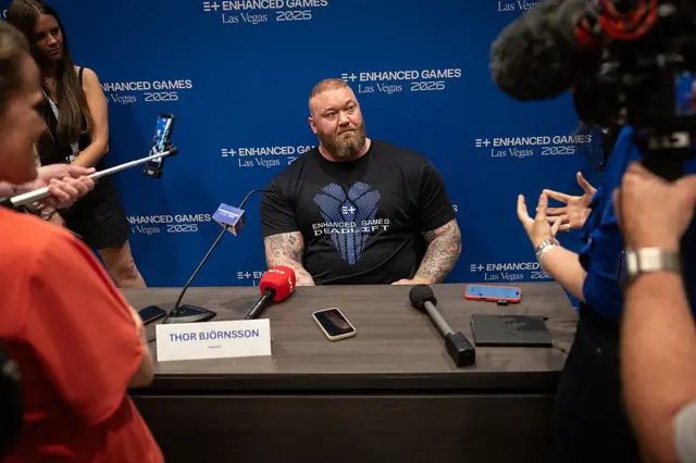 Hafthor Bjornsson siedzi przy stole konferencyjnym z mikrofonami, w tym z mikrofonem z czerwonym pokrowcem, a także z dyktafonem i dwoma smartfonami położonymi na blacie. Przed nim stoi tabliczka z napisem „THOR BJÖRNSSON”. Wokół znajdują się osoby trzymające kamery, mikrofon na wysięgniku oraz selfie stick z telefonem. W tle widoczna jest niebieska ściana z napisem „ENHANCED GAMES Las Vegas 2026”.