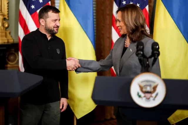 Volodymyr Zelensky e Kamala Harris dando aperto de mãos em um palco, em frente à bandeira ucraniana e à bandeira dos EUA