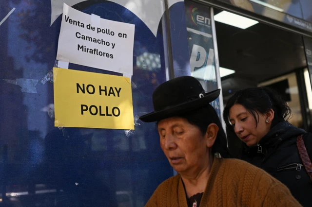 Dos mujeres caminan por el lado de una pared azul de un supermercado que tiene dos avisos: uno en fondo blanco que dice: Venta de pollo en Camacho y Miraflores y en otro en fondo amarillo que dice: No hay pollo.