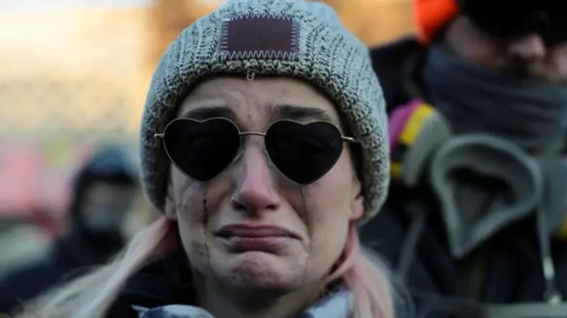 Жінка у сірій вʼязаній шапці та чорних сонцезахисних окулярах у формі серця стоїть обличчям до камери: в неї міцно стиснуті губи, а по щоках течуть чорні від туші сльози.