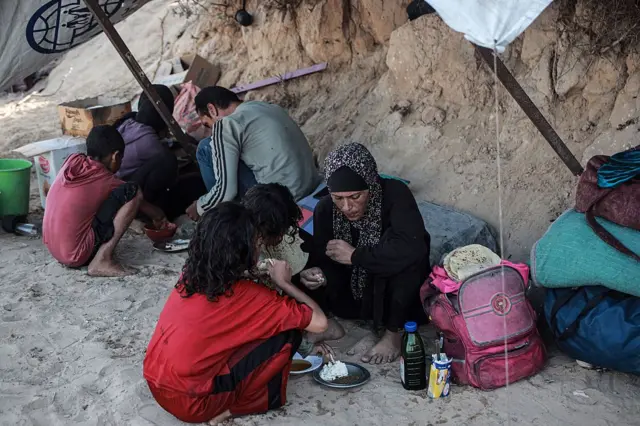 سيدة تجلس على الأرض وتضع صحناً به قطعة جبن صغيرة تأكل منها وأمامها فتاتان تأكلان من نفس الصحن، وبجوارهما حقيبة مدرسية بالية ومتهالكة.