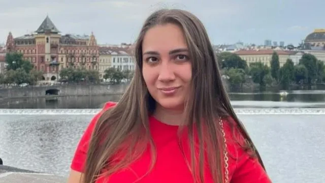 والریا دمیدنکو در لباسی قرمز رنگ کنار رودخانه‌ای که از شهر می‌گذرد ایستاده به دوربین نگاه می‌کند، پشت سر او آنسوی رودخانه ساختمان‌ها دیده می‌شوند