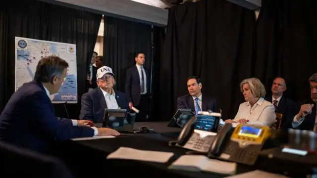 O presidente Donald Trump está sentado, usando um boné branco com a inscrição "USA", enquanto ouve o diretor da Agência Central de Inteligência (CIA), John Ratcliffe, falar. Ao redor da mesa estão o secretário de Estado dos EUA, Marco Rubio, e a chefe de gabinete da Casa Branca, Susie Wiles.