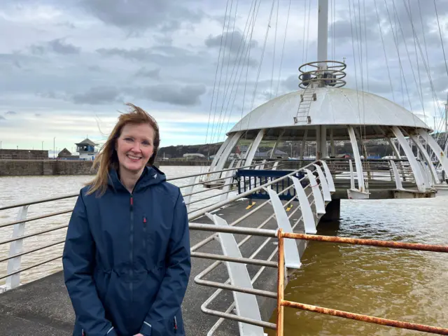 Deanne Shallcross, directora ejecutiva de la Comisión del Puerto de Whitehaven. Se encuentra en el puerto de Whitehaven, junto a la pasarela que conduce al Nido del Cuervo, una estructura blanca tipo pagoda sobre el agua. Mira a la cámara y sonríe. Tiene el pelo castaño hasta los hombros y ojos azules. Lleva una chaqueta impermeable azul.