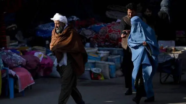 تصویر یک زن چادری پوش در شهر هرات. او در میان بازار قدم میزند. دو مرد نیز در این تصویر دیده میشوند.