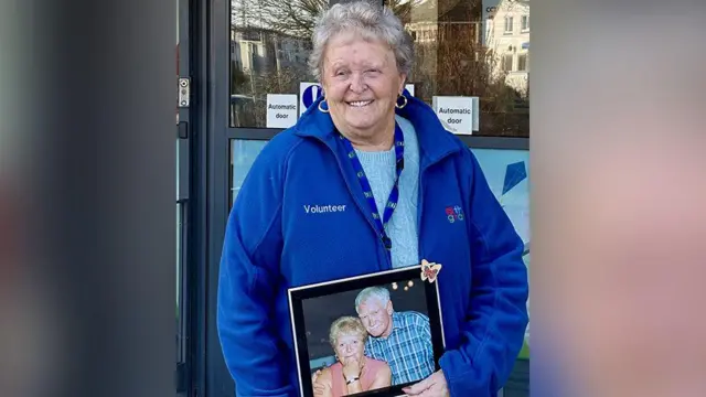 Sheila Underwood, con un polar azul y sonriendo, sostiene una fotografía enmarcada de ella con su difunto esposo Jim frente al centro comunitario Grange. Jim lleva una camisa azul a cuadros y abraza a Sheila, quien lleva una blusa color melocotón sin mangas. Ambos sonríen.