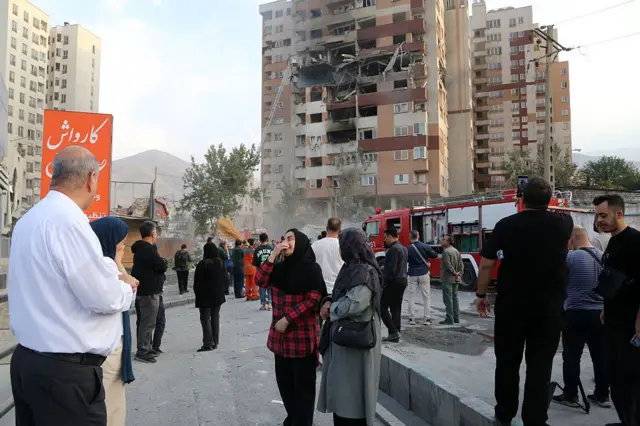 یک ساختمان آسیب‌دیده و مردمی که بیرون آن ایستاده‌اند، امدادرسانان در حال کار هستند