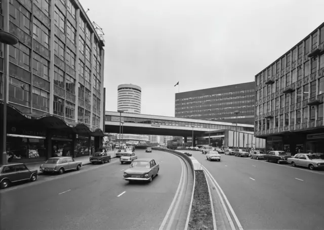 Imagen en blanco y negro del Ringway Centre de Birmingham en la década de 1970, con coches circulando por la amplia vía. La rotonda de la ciudad se ve al fondo con coches aparcados a ambos lados de la carretera.