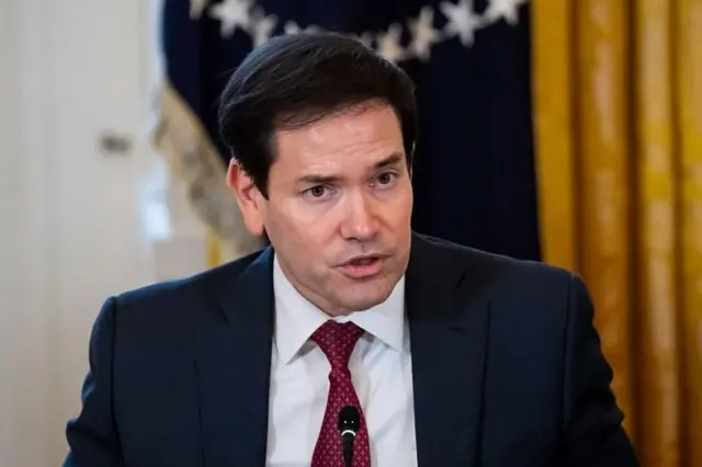 Rubio resmi giyimli bir mikrofona konuşuyor, arka planda sarı siyah perdeler var. 