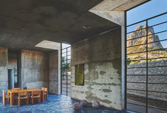 Interior de arquitectura de hormigón visto con mesa de madera y grandes ventanales que enmarcan un paisaje rocoso.