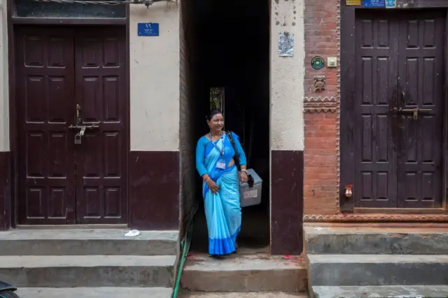 नीलो सारी लगाएर खोपको डिब्बा बोकेर घरबाट बाहिर निस्किरहेकी एक जना महिला स्वास्थ्य स्वयंसेवी। 