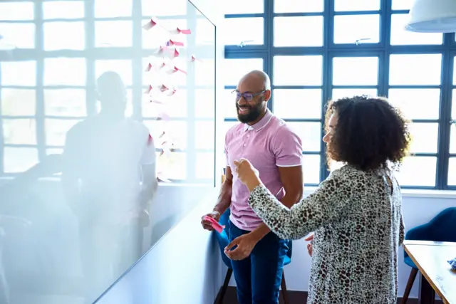 Duas pessoas sorrindo em frente a lousa, onde colocam anotações em post-it