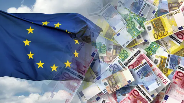 Zdjęcie ilustracyjne przedstawiające flagę Unii Europejskiej, która jest niebieska z żółtymi gwiazdkami ułożonymi w kółko, oraz rozsypane banknoty euro