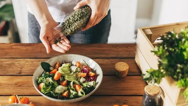 Тарілка із салатом стоїть на столі. Жіноча рука додає туди насіння гарбуза