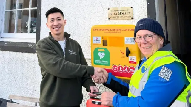 El Sr. Morley le estrecha la mano a un hombre de Olive Social Coffee House. Sonríen frente al desfibrilador amarillo brillante colgado en la pared. Sobre él, una placa dice: "En memoria de Billy Morley".