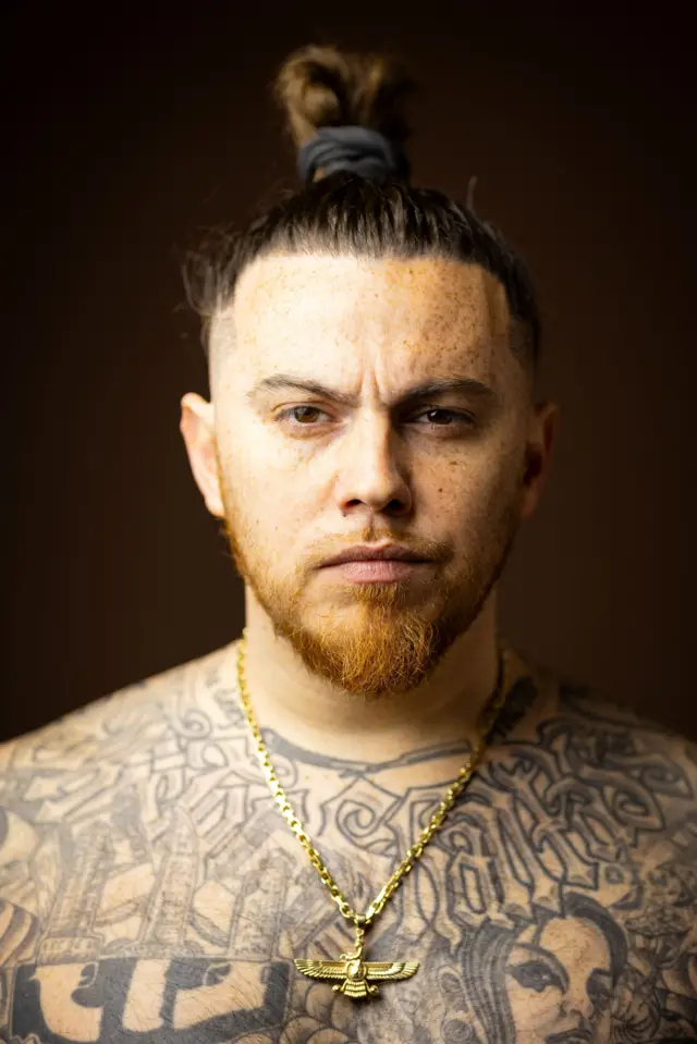 Retrato de estudio en primer plano de una persona con el cabello recogido en un moño, pecas, barba corta y numerosos tatuajes en blanco y negro en el pecho y los hombros. 