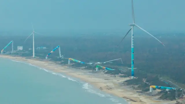 Una playa con una fila de seis turbinas eólicas, cuatro de ellas dobladas y caídas sobre el suelo 