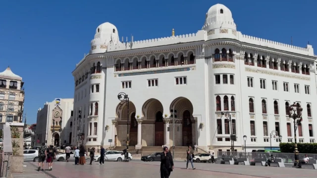 White mosque dey stand against clear blue sky as pedestrians dey walk along one busy street and cars dey pass.