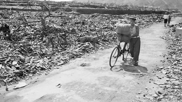 A Japanese civilian pushes his loaded bike down a path which has been cleared of rubble in the aftermath of the 9 August 1945 attack. On either side of the path debris, twisted metal, and gnarled tree stumps fill the area