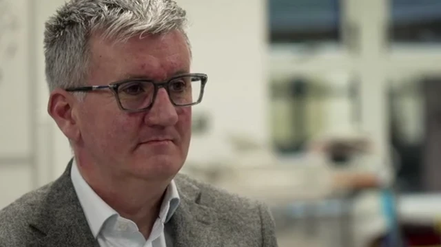 Se ve a Matthew Trainer, con gafas y una chaqueta, sentado en el interior, mirando ligeramente hacia un lado.