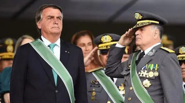 Bolsonaro em pé, com olhar solene, enquanto Freire Gomes, ao seu lado, presta continência