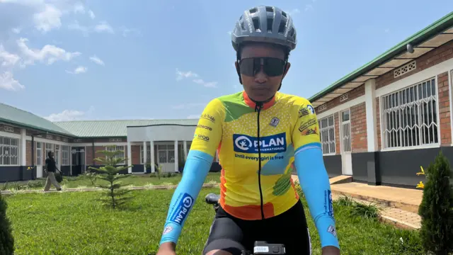 Olivia Manigragena yambaye ingofero yo kwirinda y'ikijuju, amataratara y'izuba, n'umwenda w'umuhondo n'ubururu yicaye ku igare areba muri camera mbere y'isiganwa ku kigo cya Bikes for Future kiri mu Bugesera. 