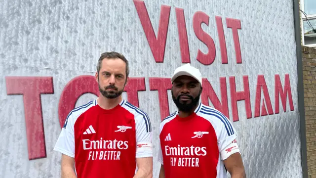 James Turner et Tresor Kubadika portent des maillots rouge et blanc d'Arsenal et des brassards « Gunners for Peace » devant un panneau d'affichage portant le message « Visit Tottenham » (Visitez Tottenham).