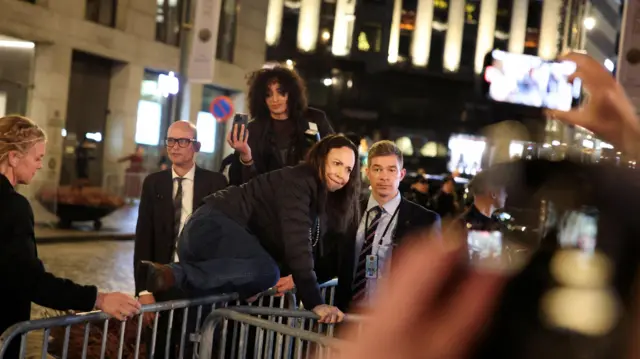 María Corina Machado salta as grades de segurança diante do Grand Hotel, em Oslo, para cumprimentar apoiadores que aplaudem, enquanto seguranças observam