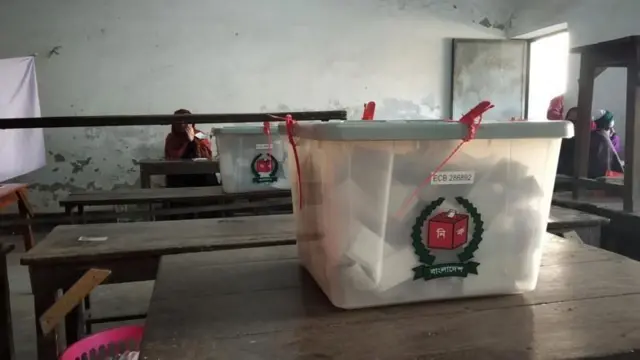 একটি টেবিলের ওপর ব্যালট ভর্তি ভোটের বাক্স রাখা। পেছনে আরেকটি বেঞ্চের ওপর একই ধরনের আরেকটি বাক্স। তার পেছনে একটি বেঞ্চে বসে আছেন একজন নারী 