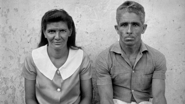 Dona Inês e Seu Mundinho, pais de João Mendes, posam sentados em frente a uma parede