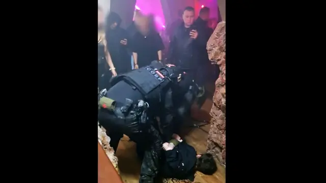 Członek rosyjskich organów ścigania pochyla się nad osobą leżącą na podłodze imprezy, trzymając ręce za plecami, podczas gdy inni goście obserwują. Klub nocny ma różowe neonowe oświetlenie.