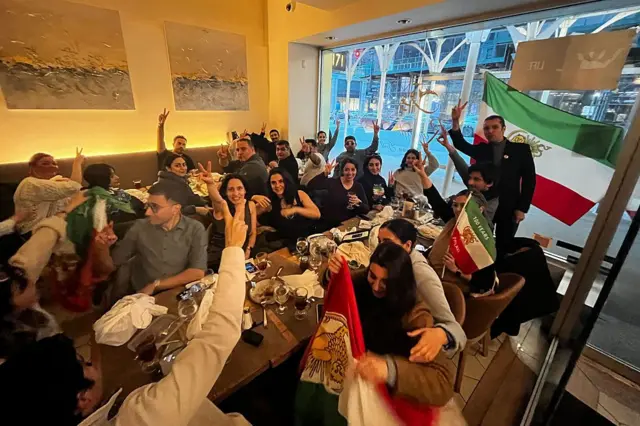 1 Mart 2026 Pazar günü, New York'un Midtown semtindeki Ravagh Persian Grill restoranında müşteriler, İran'ın Dini Lideri Ayetullah Ali Hamaney'in vefatını kutluyor. Fonda İran bayrağı ve onun önünde eğlenen kalabalık bir grup görülüyor. 