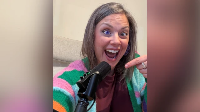 Una mujer de cabello gris y castaño sonríe y señala con la mano un micrófono negro para podcast. Lleva un cárdigan naranja, rosa, verde y azul, y tiene un ojo morado.