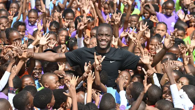 Letsile Tebogo, vêtu d'un T-shirt noir, est assailli par des centaines d'enfants souriants, dont beaucoup ont les mains levées vers lui.