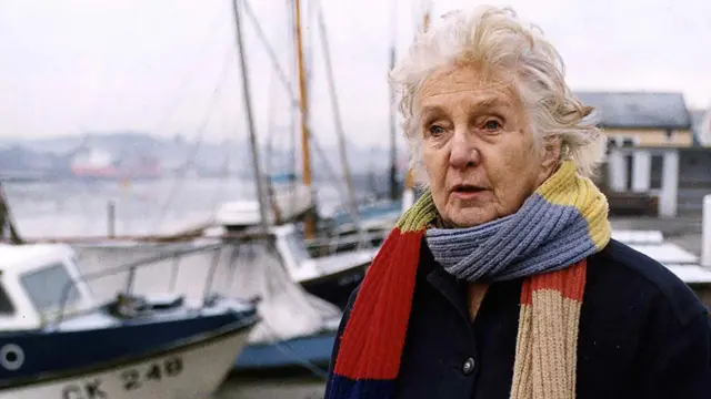 Starija žena svetlih očiju sa šarenim šalom oko vrata, iza nje su brodovi