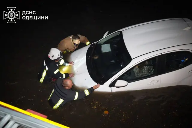 Стоячи по пояс у воді, рятувальники штовхають білу автівку, у якій сидить людина в леопардовій кофті