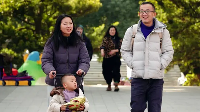 Mor renkli şişme mont giymiş bir anne ve beyaz renkli şişme mont giymiş bir baba, bebek arabasında küçük bir çocukla parkta dolaşıyorlar.