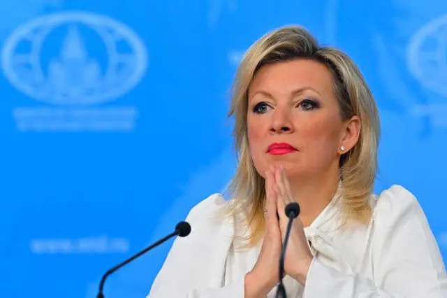 Zakharova mavi bir fon önünde beyaz gömlekli, mikrofonların önünde oturuyor, ellerini yüzünün altında birleştirmiş.